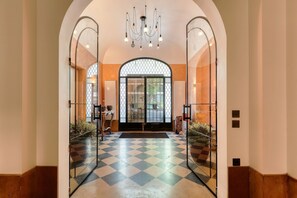 Eine Hotelhalle mit Schachbrettmuster-Boden, gespiegelten Wänden, einer Kristallleuchtreibe und einem großen Rundbogenportal.