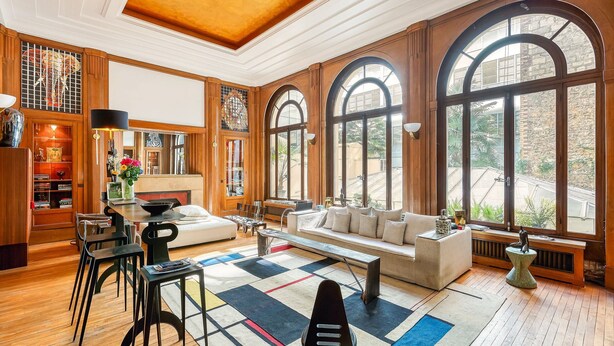 A spacious living area with a wooden floor, a sofa, a coffee table, and a dining table with chairs. Large arched windows provide natural light and a view of the outdoors. - Par056 - Mansion-Style Apartment by Parc Monceau (Paris)