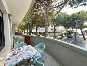 A balcony with a table set for two, chairs, and a view of the street and buildings. - Familiar Apartment Close to the Beach (Lignano Sabbiadoro)