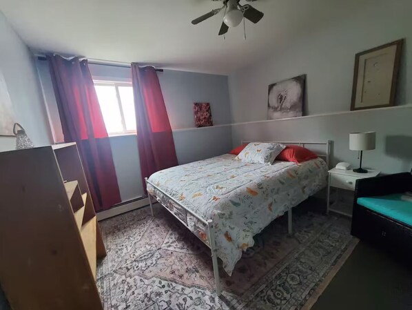 A bedroom with a bed, a nightstand, a desk, a chair, a ceiling fan, and a window with curtains.