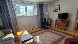Una sala de estar con sofá, mesa de centro, televisor, aparador de madera y ventana con cortinas.