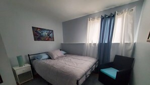 Un dormitorio con cama, una silla, una mesita de noche, un cuadro y una ventana con cortinas.