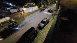 Et nattetidsbillede af en gade med parkerede biler, en bygning med skiltet 'SCHOOL ASIAN CENTRE', og en murstensmur til højre.