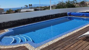 Piscina com deck de madeira, rodeada por um muro de pedra, e vista clara do oceano e montanhas ao fundo.