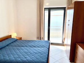 Et hotelværelse med seng, et natbord, en lampe, et klædeskab og en skydedør, der fører ud til en balkon.