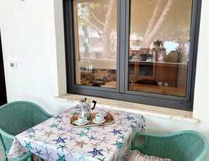 Ein kleiner Tisch mit Tischdecke in Form eines Seesterns, zwei grüne Stühle und ein Fenster mit Blick auf Gebäude und Bäume.