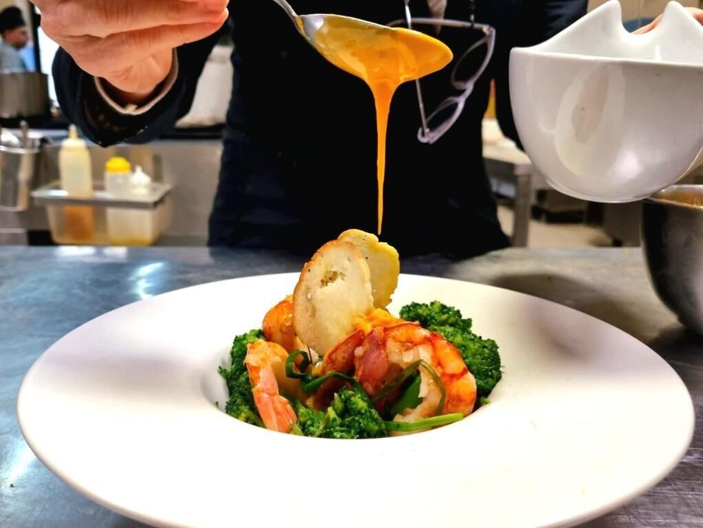 A person pouring sauce over a dish with shrimp and broccoli on a white plate.