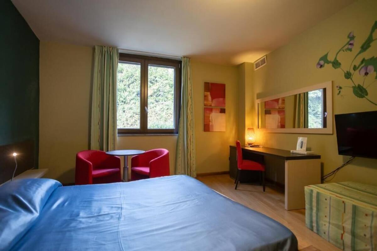 A hotel room with a bed, a desk, two red chairs, a television, and a window with curtains.