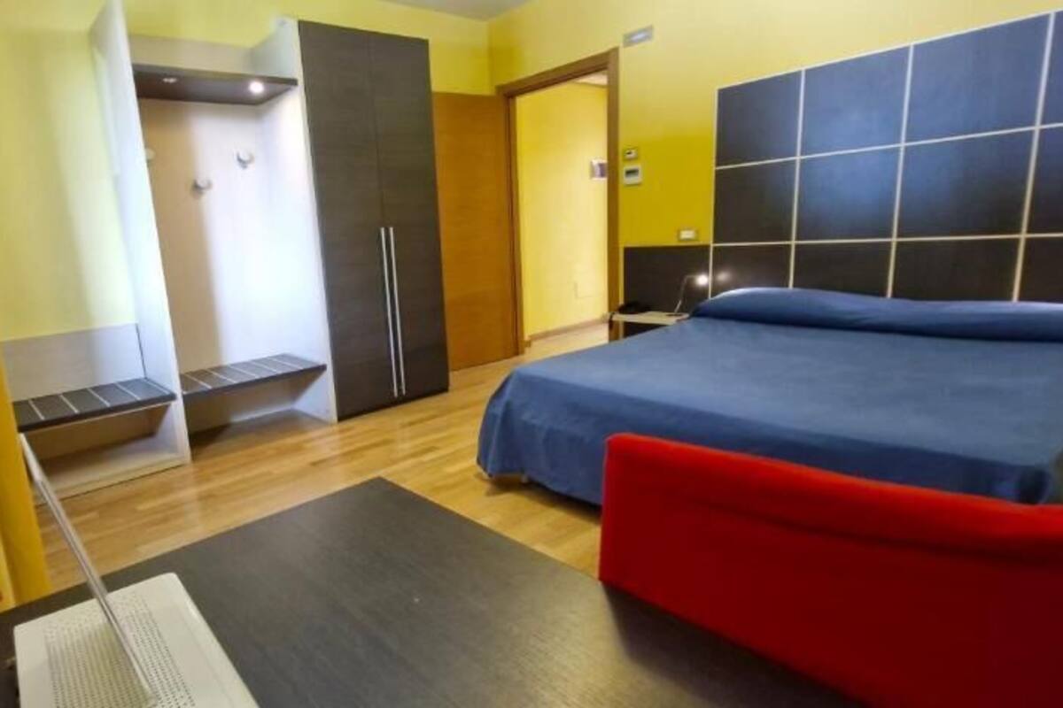 A hotel room with a bed, a red chair, a wardrobe, and a desk with a chair.