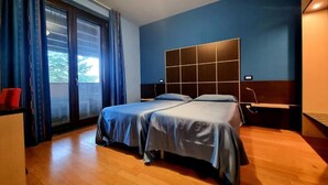 Ein Hotelzimmer mit zwei Betten, einem Schreibtisch, einem Stuhl, einem Fenster mit Vorhängen und Blick auf Bäume.