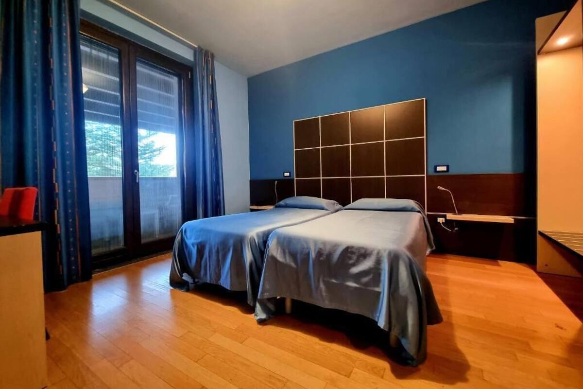 A hotel room with two beds, a desk, a chair, a window with curtains, and a view of trees.