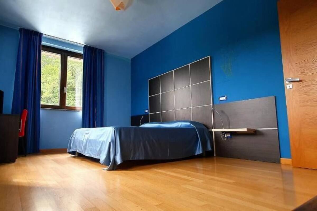 A bedroom with a bed, a window with curtains, a wooden floor, and a door.