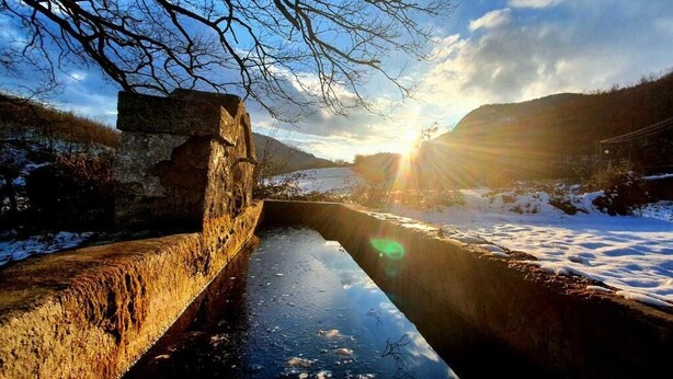 Eine Steinbrücke über einem zugefrorenen Fluss, wobei sich das Sonnenlicht im Wasser spiegelt.