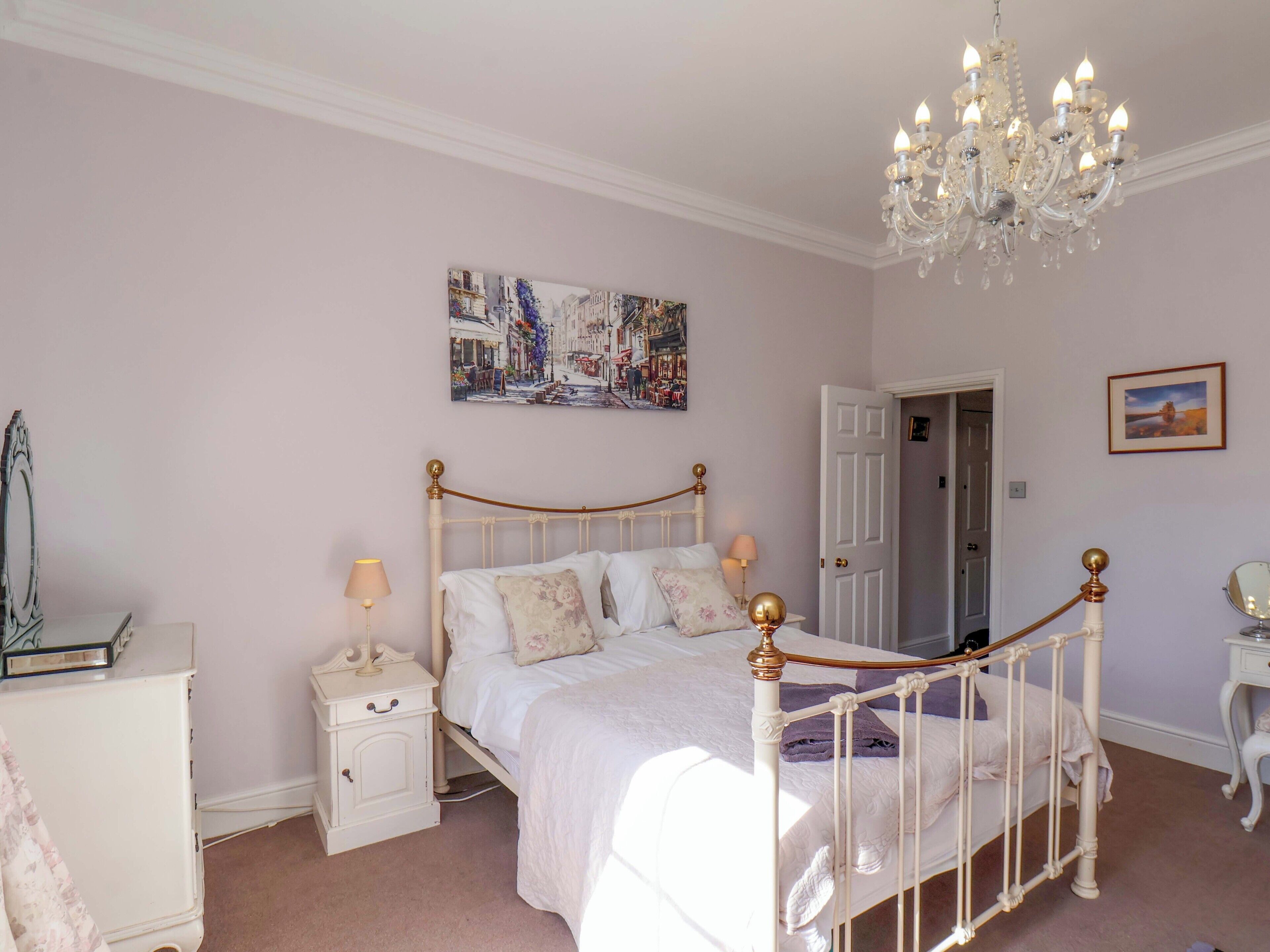 A bedroom with a white bed, a nightstand, a dresser, a chandelier, and a door.