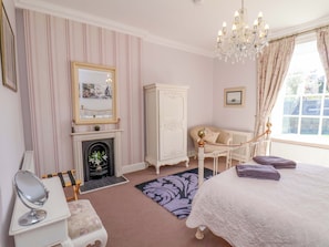 A bedroom with a fireplace, a bed, a chair, a mirror, and a chandelier. - Annie's Place (Whitby)