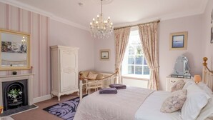Ein Schlafzimmer mit Bett, Kommode, Stuhl, Spiegel, Kamin und einem Fenster mit Vorhängen.