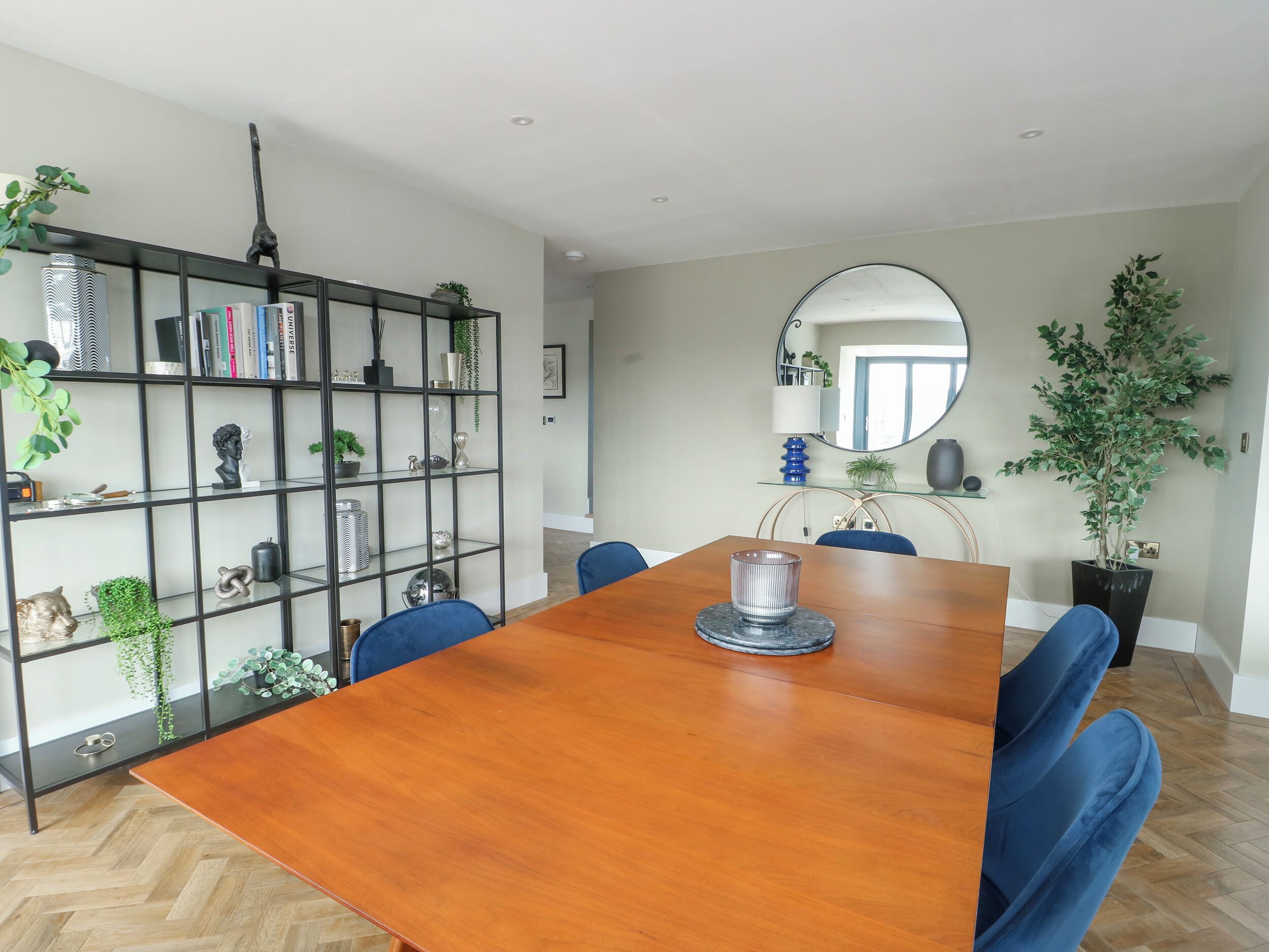 Un comedor con una mesa de madera, sillas azules, una estantería de metal negro y un espejo en la pared.