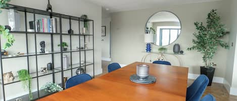 A dining area with a wooden table, blue chairs, a black metal bookshelf, and a mirror on the wall.