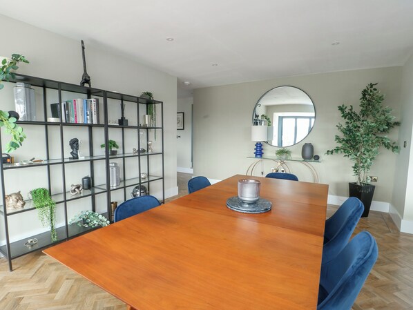 Un comedor con una mesa de madera, sillas azules, una estantería de metal negro y un espejo en la pared.