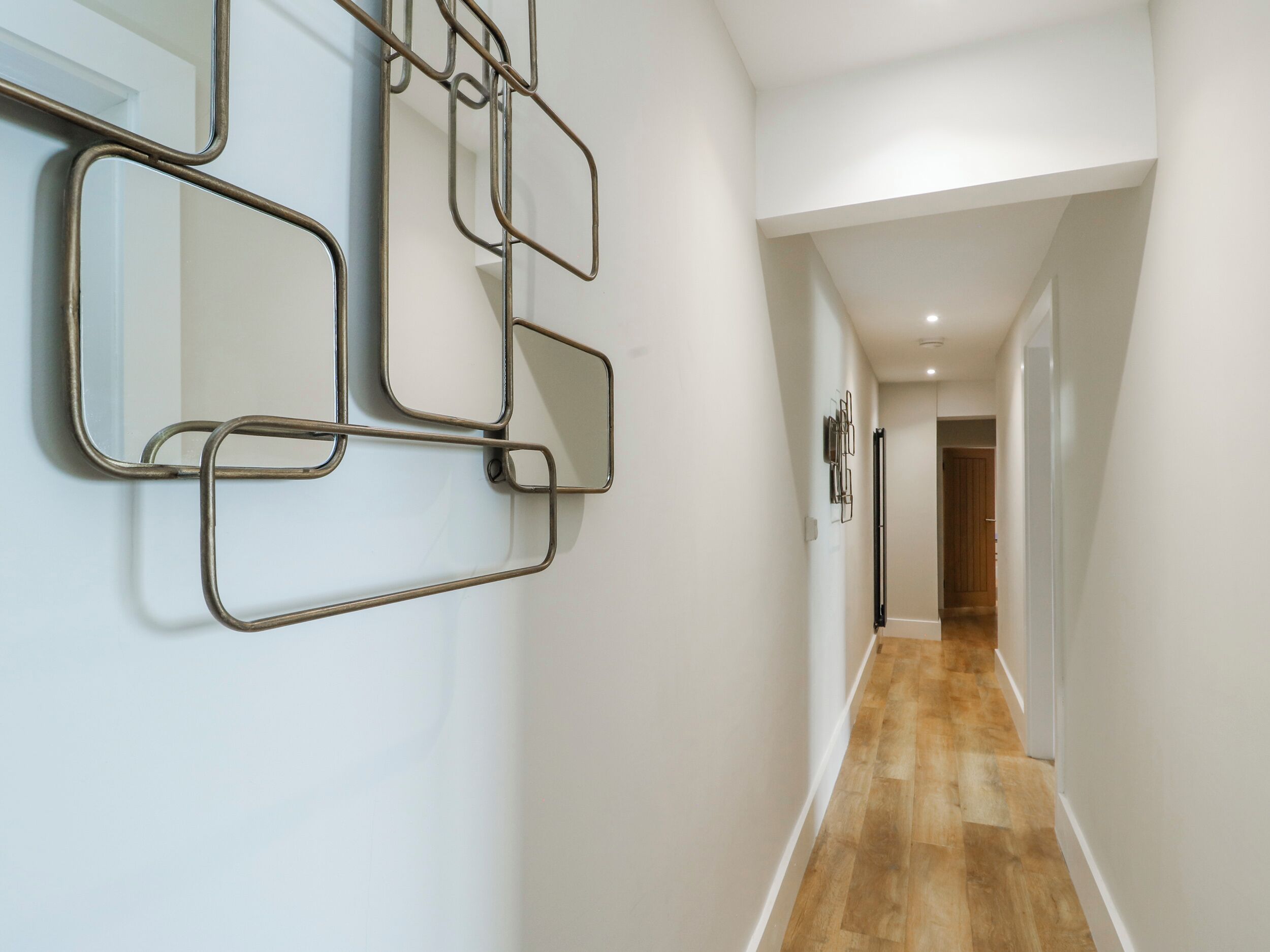 A hallway with a wooden floor, white walls, and a wall-mounted mirror with a geometric metal frame.