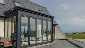 A modern conservatory with a glass enclosure, a seating area with a chair and table, and a view of the landscape. - Pengarnedd (Amlwch)