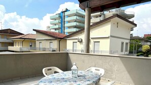 A balcony with a table and chairs, a white lantern, and a building with balconies in the background. - Great Accommodation for Groups in Lignano (Lignano Sabbiadoro)