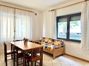 A room with a dining table, chairs, a sofa, a window with curtains, and a view of the outside. - Great Accommodation for Groups in Lignano (Lignano Sabbiadoro)