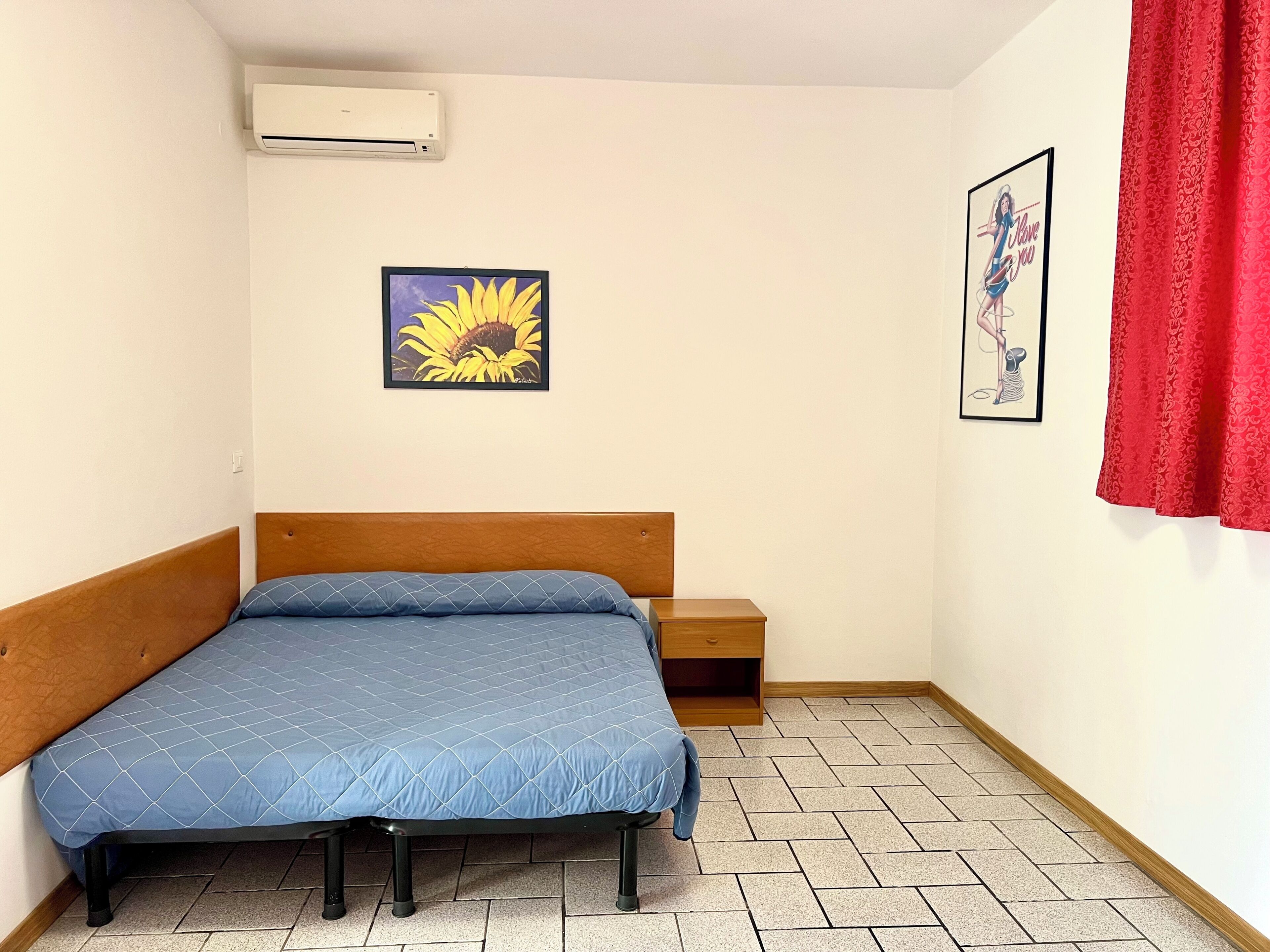 Una camera piccola con un letto singolo, una testiera in legno, una coperta trapuntata blu, un comodino, un condizionatore d'aria, un quadro con girasole e un poster incorniciati appesi al muro.