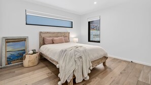 A bedroom with a wooden bed, a nightstand, a bedside table, a lamp, a framed painting, and a large window with a view. - 9 Oceania (Lorne)