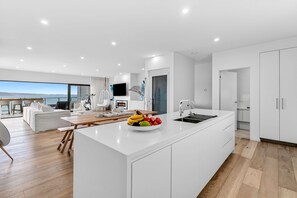 A modern kitchen with a central island, a dining area with a view, and a living space with a sofa and chairs. - 9 Oceania (Lorne)