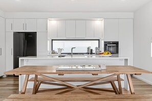 A modern kitchen with white cabinetry, a central island with a sink, and a wooden dining table. - 9 Oceania (Lorne)
