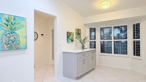 Una cocina luminosa con gabinetes blancos, una encimera y una ventana con persianas. Hay dos cuadros en la pared y una lámpara en el techo.