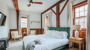 Une chambre à coucher comprenant un lit, une tête de lit en bois, une table de chevet, une lampe, une chaise, une fenêtre et un ventilateur de plafond.