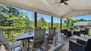 A covered outdoor deck with a table and chairs, surrounded by a railing, overlooking a wooded area.