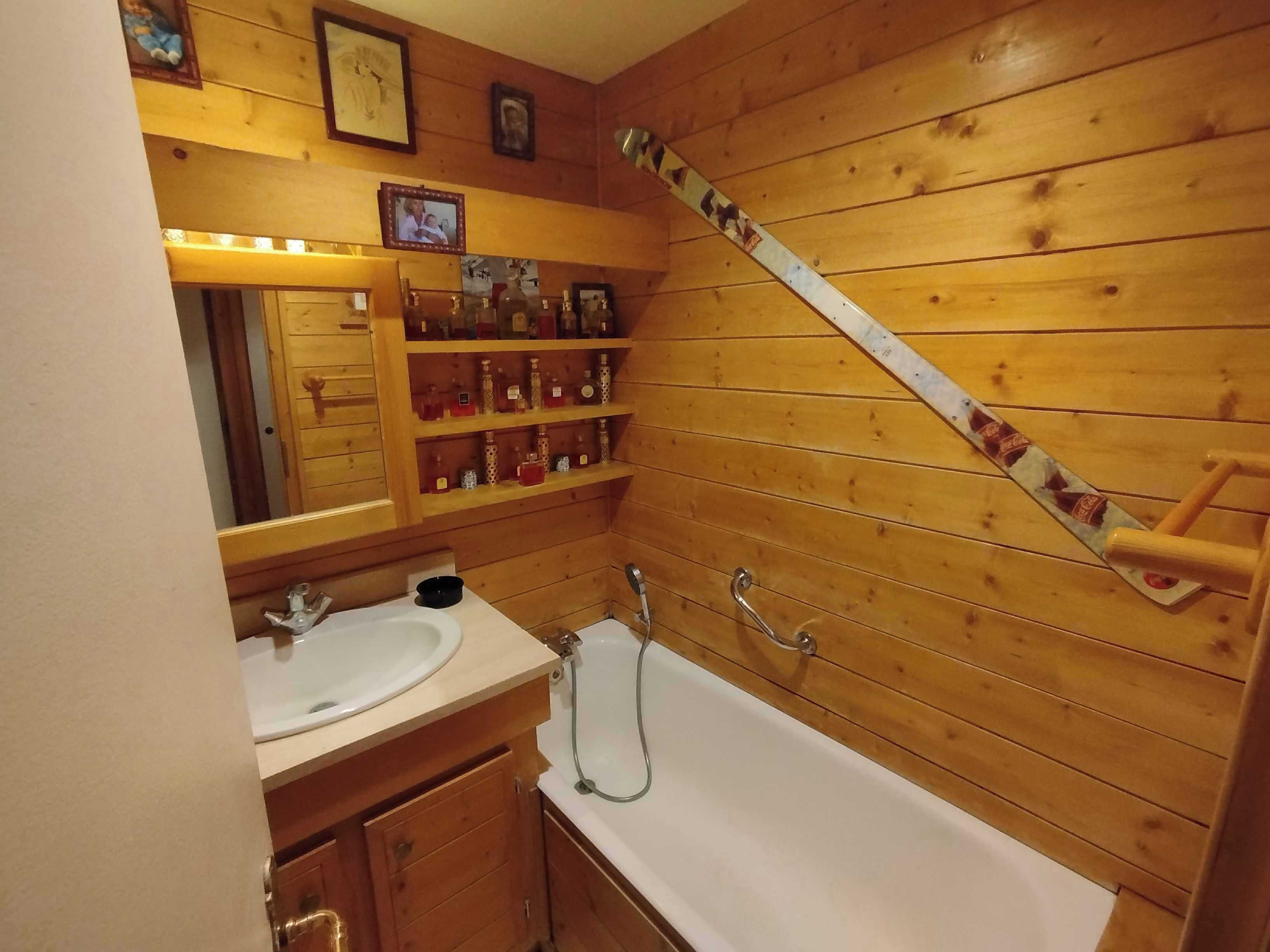 A wooden-paneled bathroom with a sink, mirror, shelves with bottles, and a shower.