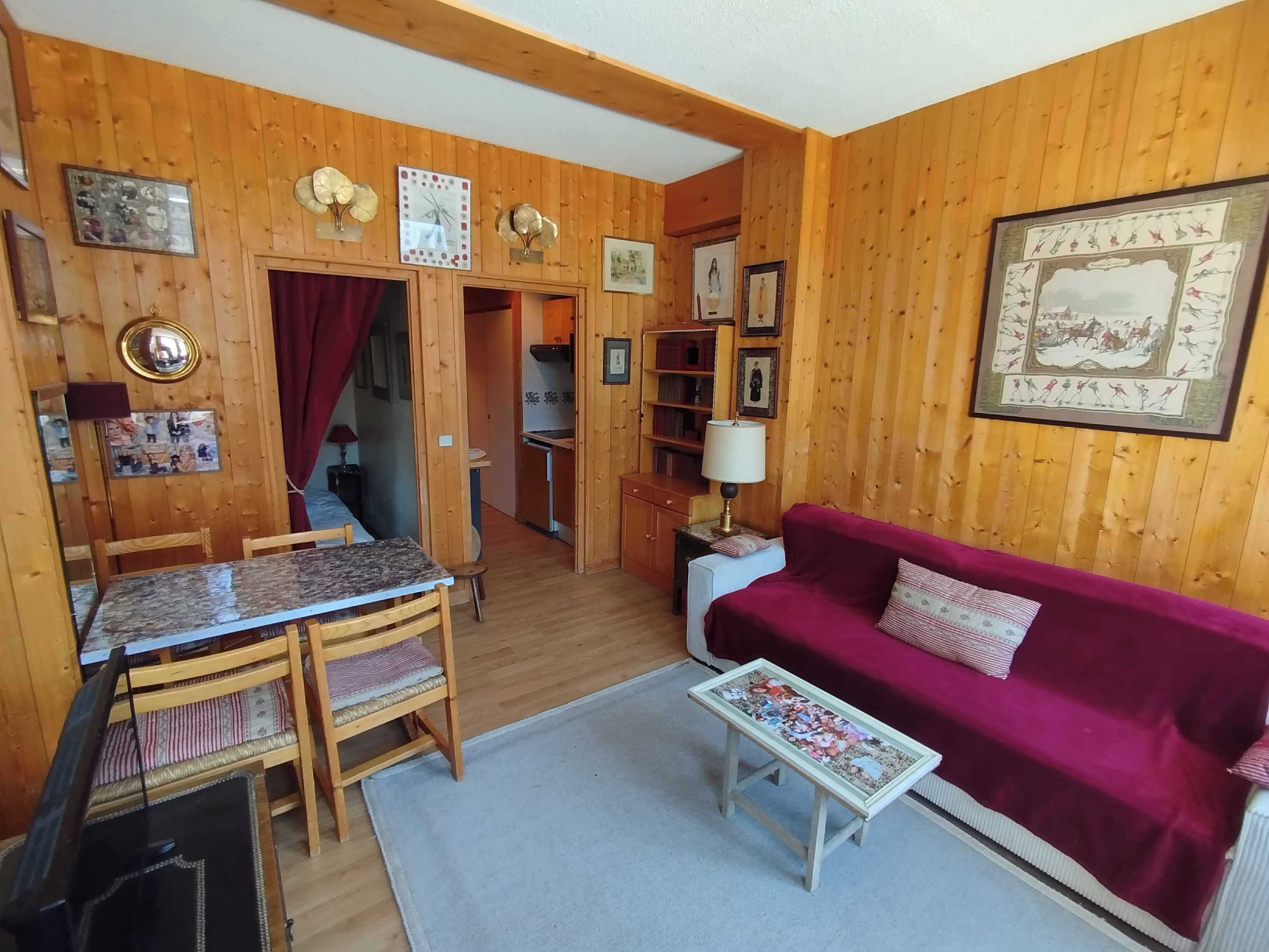 Una habitación con paneles de madera, una mesa de comedor, sillas, un sofá rojo, una mesita de centro y una zona de cocina al fondo.