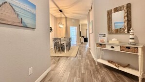 Um corredor com piso de madeira, área de jantar com mesa e cadeiras, mesa de consola com espelho e objetos decorativos, e um quadro na parede.