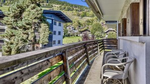 Un balcone con ringhiera in legno, sedie in vimini e vista su una zona montuosa con case.