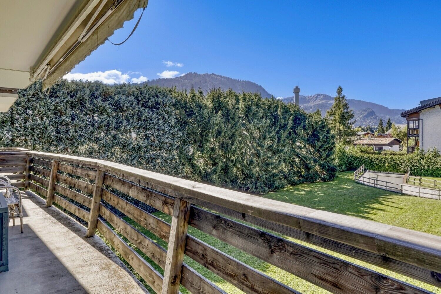 A balcony with a wooden railing overlooking a mountainous landscape, a grassy area, and a building with a balcony.