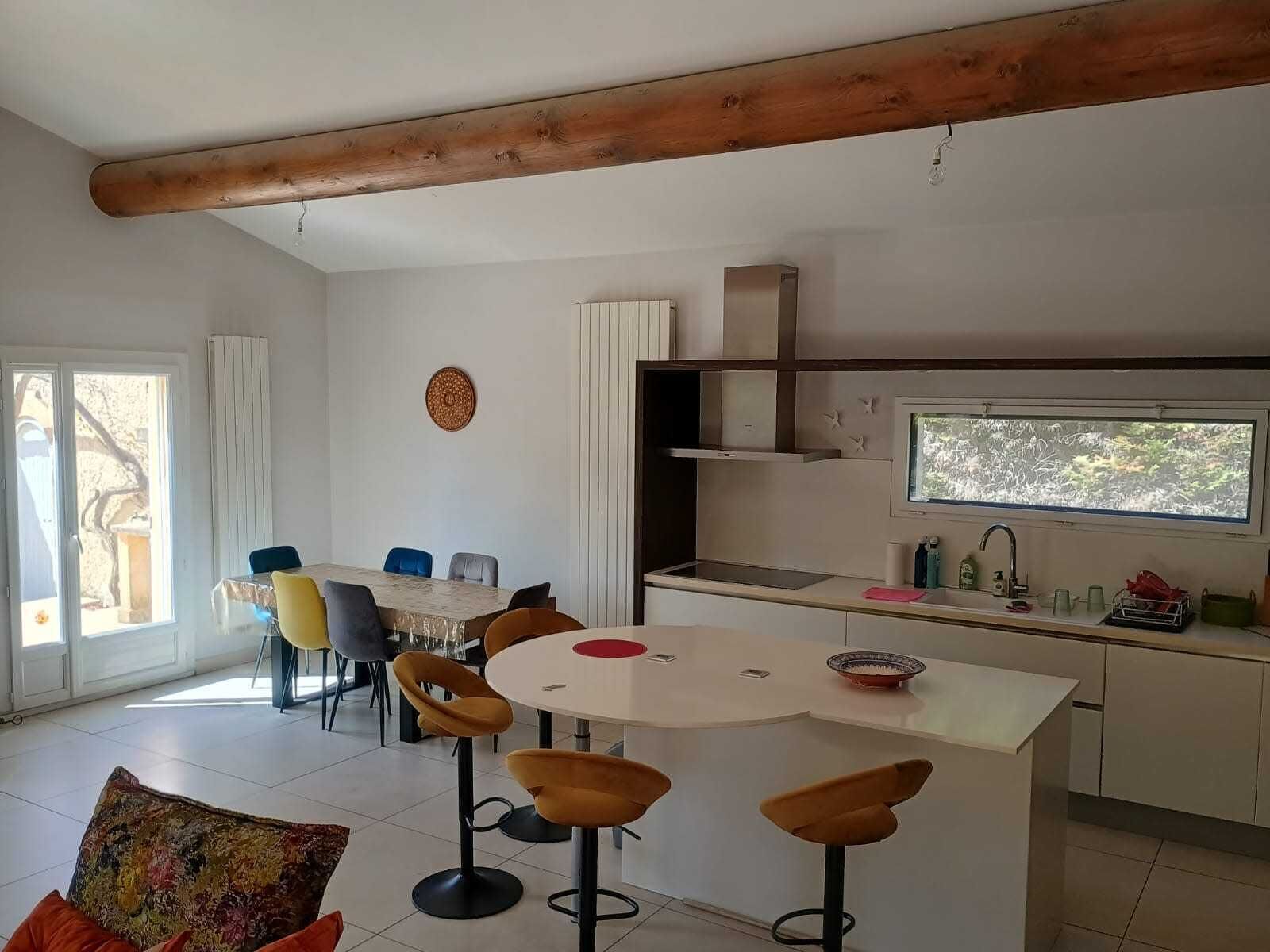 Une cuisine moderne avec un coin repas, des poutres en bois au plafond, une grande fenêtre et une vue sur la verdure.