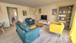 Ein Wohnzimmer mit Sofa, Sesseln, Couchtisch, Fernseher, Bücherregal und einer Tür zu einem anderen Raum.