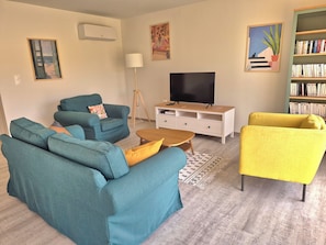 Un salon avec des canapés de couleur turquoise, un fauteuil jaune, un téléviseur, une bibliothèque et un tableau représentant un chat.