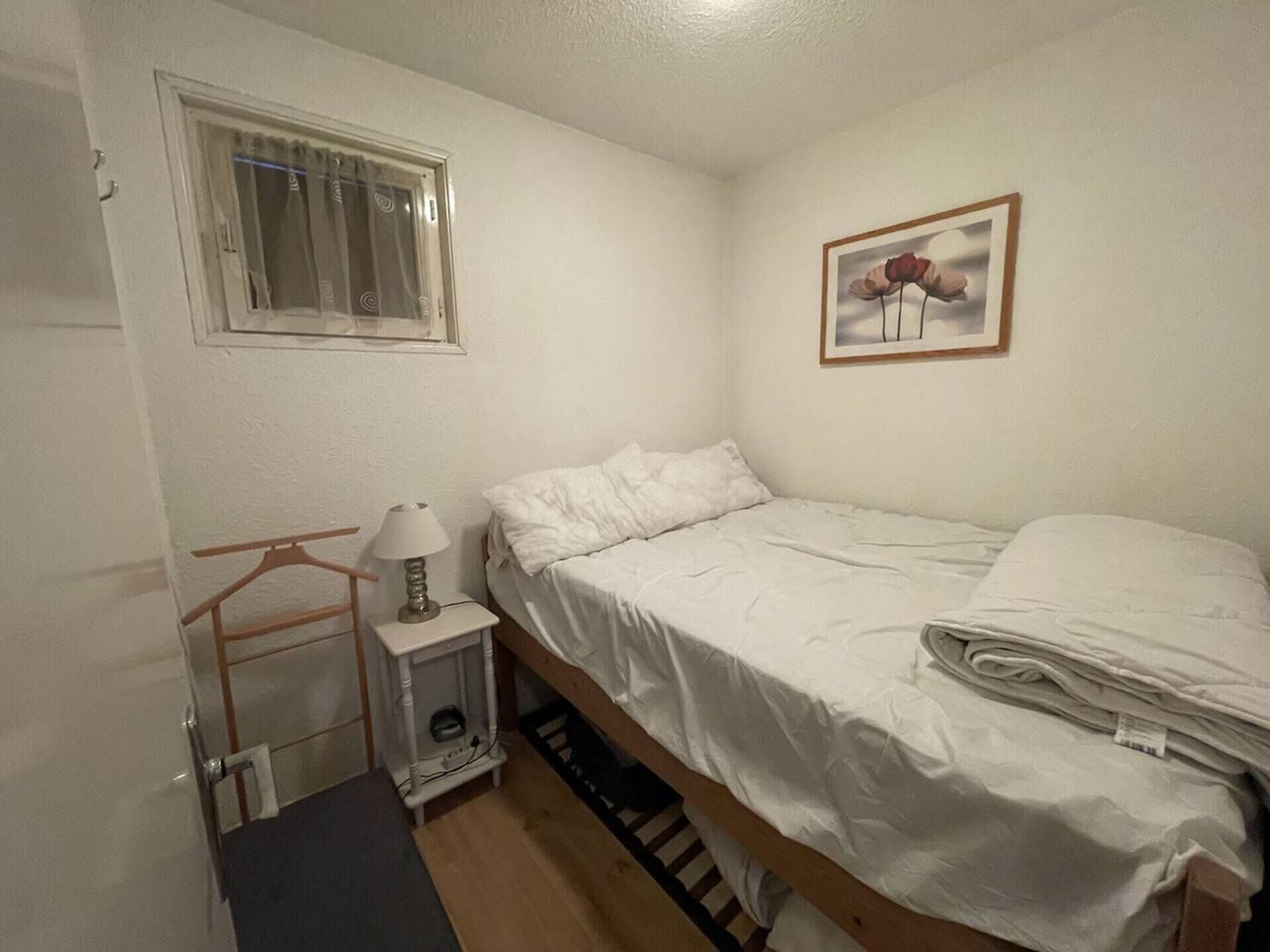 Une petite chambre avec un lit simple, une fenêtre, une table de chevet avec une lampe, un tableau encadré au mur et un plancher en bois.