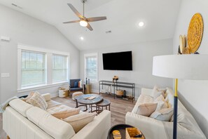 A spacious living room with a ceiling fan, a flat-screen TV mounted on the wall, and a large window with blinds. - New, dog-friendly 3BR with deck & sound view (Kill Devil Hills)