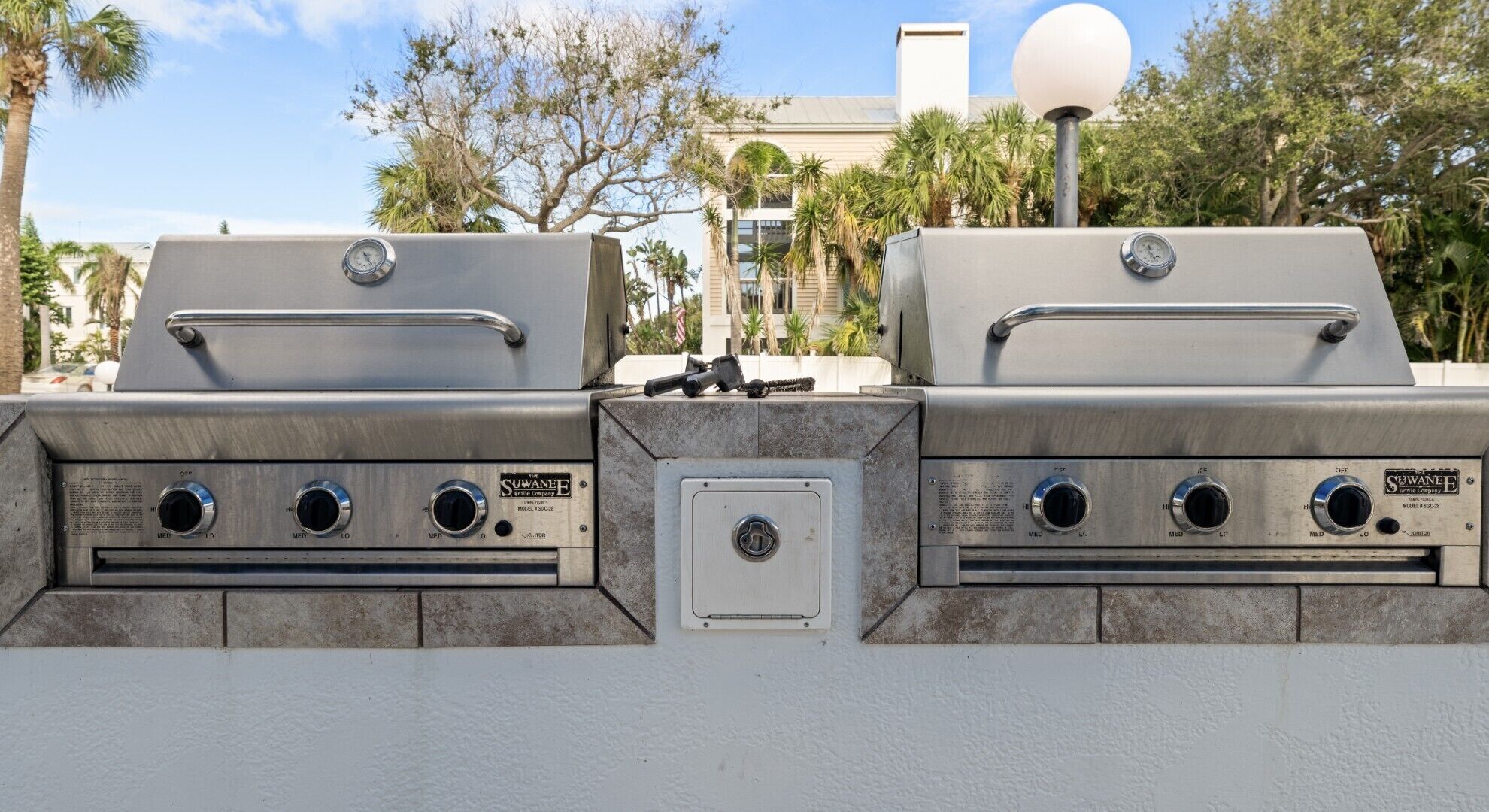 A two-burner stainless steel grill with control knobs and a thermometer, mounted on a white wall with a small electrical box below.