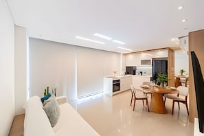 A modern living room with a sofa, dining table, and chairs, a kitchen area with appliances, and a large window providing natural light.