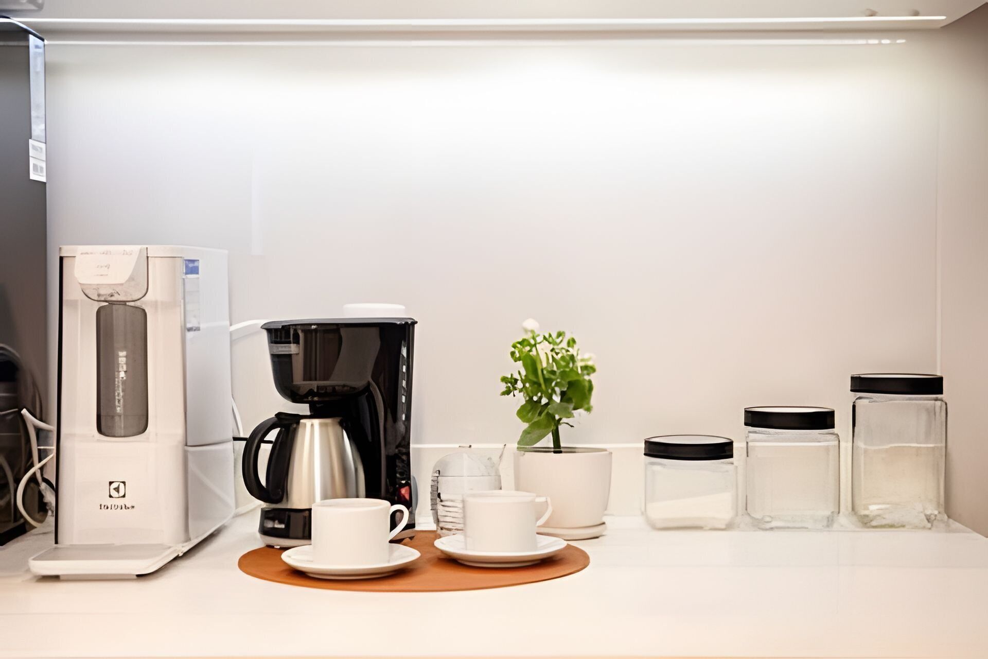 Una kitchenette con cafetera, dos tazas, una pequeña planta en maceta y frascos de almacenamiento.