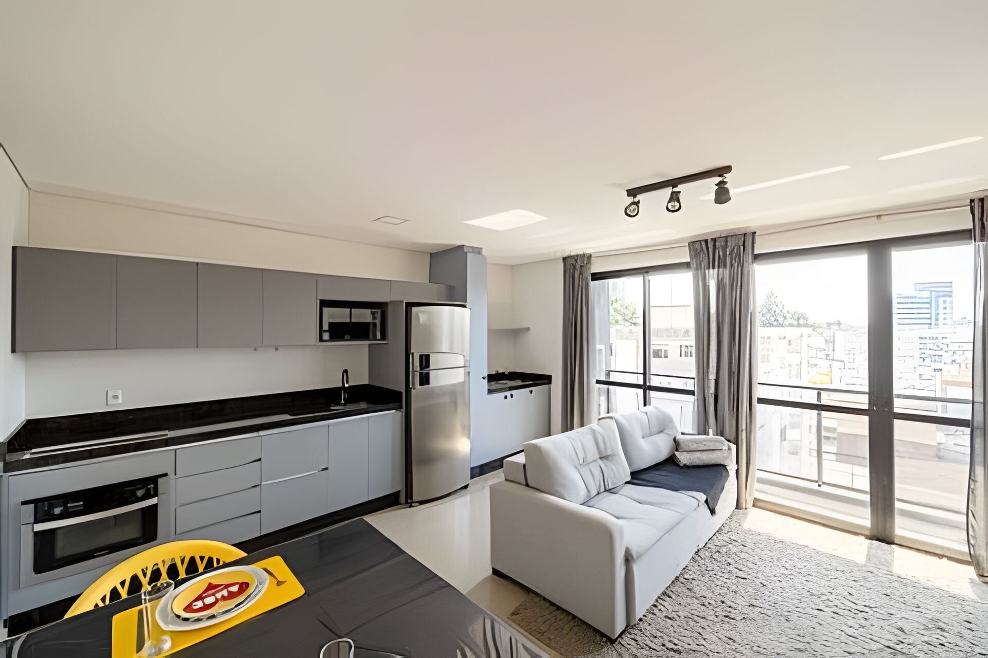 Cocina moderna con gabinetes grises, encimera negra y refrigerador de acero inoxidable. Hay un sofá gris y una mesa de comedor con un mantel individual amarillo. Amplios ventanales ofrecen vistas de la ciudad.