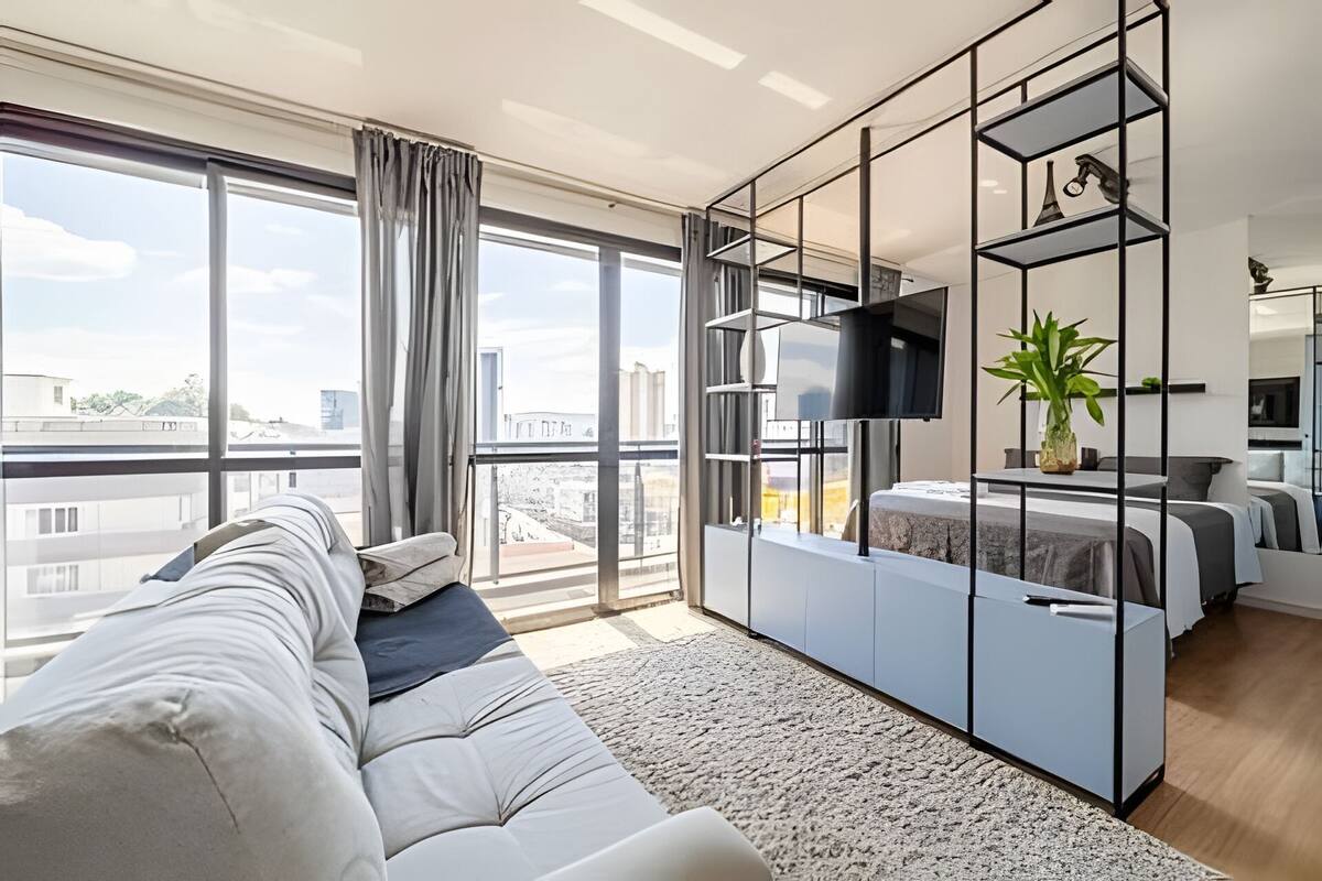 A modern living room with a sofa, a TV mounted on a wall, and a balcony with a view of the city.