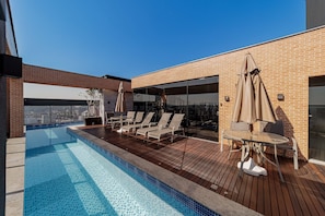 A rooftop pool with lounge chairs and umbrellas, a wooden deck, and a brick building with large windows.